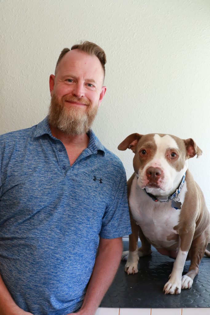 a person standing next to a brown and white pitbull breed dog. Person is wearing a blue shirt and has a beard with gray and red.